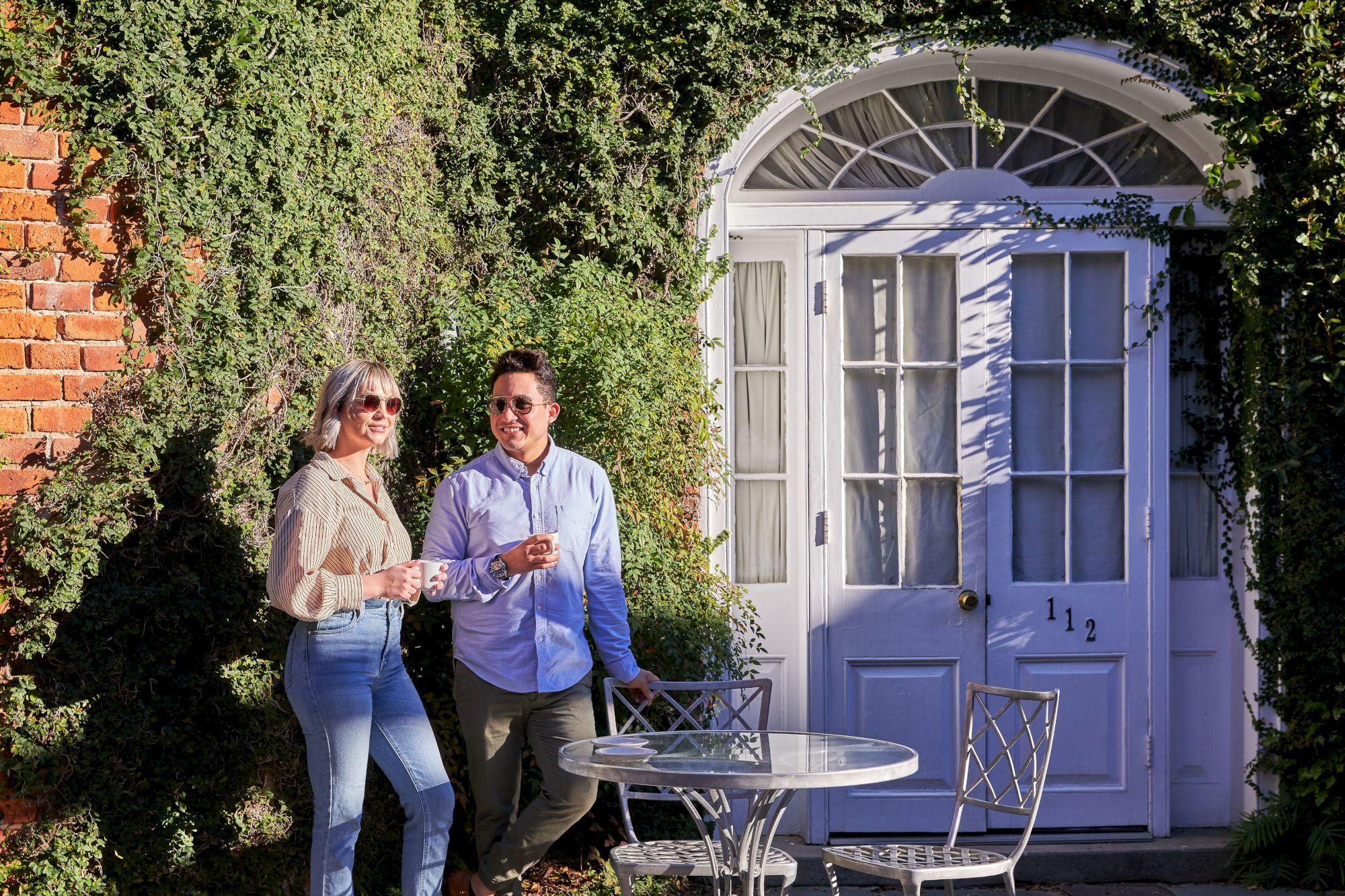 A couple stands smiling near a vine-covered wall and door, holding mugs near a metal table with chairs in a sunny setting.