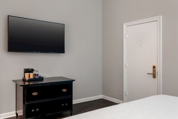 A hotel room corner with a wall-mounted TV, dresser with coffee maker, and a door. White walls and minimal decor create a calm space.