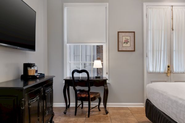 A room features a bed, TV, ornate desk with chair, a lamp, coffee maker, and framed art, with soft lighting and neutral decor.