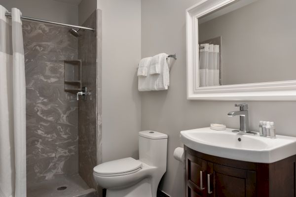 A modern bathroom with a shower, toilet, sink, and mirror. Towels are neatly hung on a rack, and the decor is minimalist.