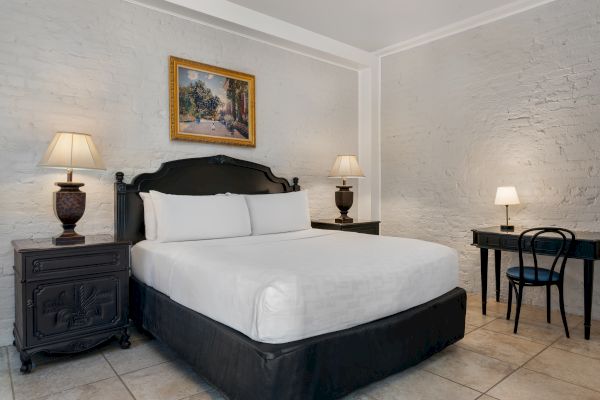 A cozy bedroom with a bed, two lamps on nightstands, a painting above, and a desk with a chair and small lamp on the right side.