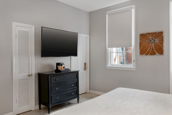 A bedroom with a TV, dresser, and coffee maker. Artwork on the wall and a window with a blind complete the decor.