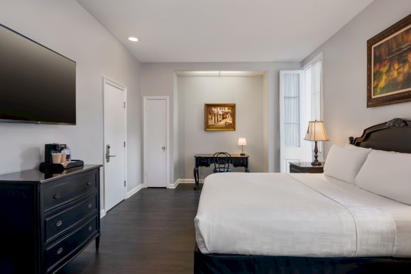 A modern hotel room with a large bed, wall-mounted TV, desk, lamps, and artwork, featuring neutral tones and minimalistic decor.