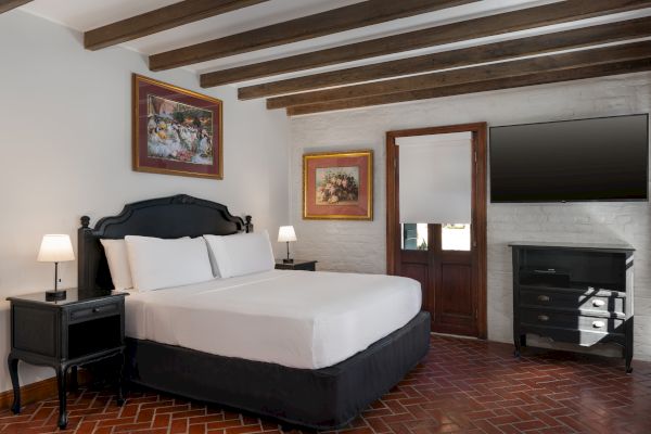 A cozy bedroom with white walls, featuring a black-framed bed, bedside lamps, paintings, wooden beams, a TV, and brick flooring.