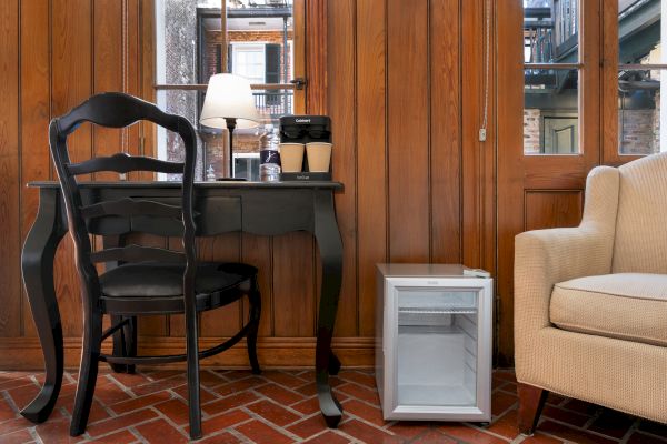 The image shows a black desk with a chair, a table lamp, coffee cups, a beige armchair, and a small fridge on a brick floor.