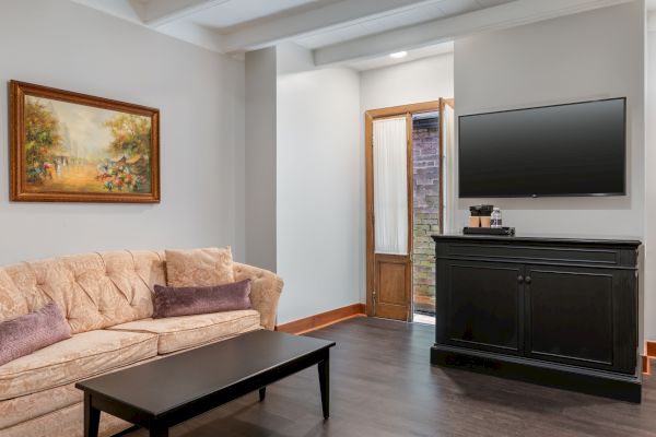 A living room with a beige sofa, black coffee table, wall-mounted TV, wooden cabinet, painting, and an open door.