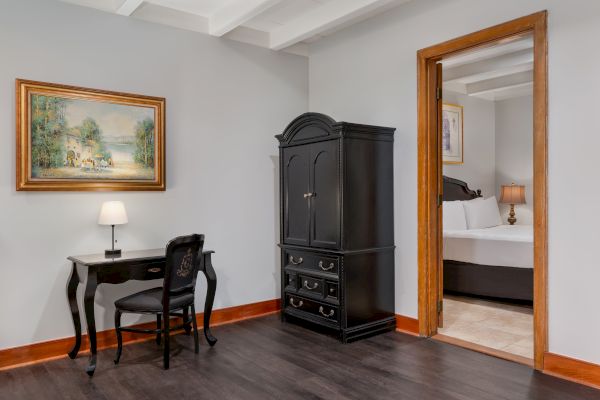 A room with a small black desk, chair, lamp, black armoire, painting, wood trim, and an open door to a bedroom with a bed and lamp.