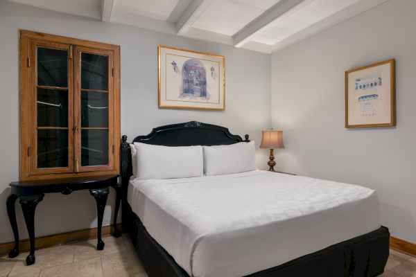 The image shows a cozy bedroom with a double bed, two artworks on the walls, a wooden cabinet, and a lamp on a small table.