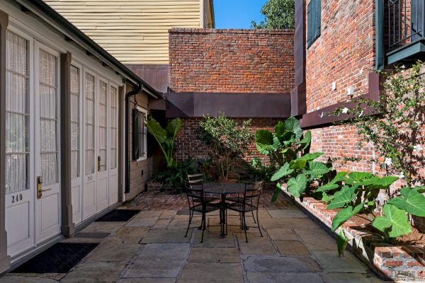 A cozy patio area with a metal table and chairs, surrounded by brick walls and lush greenery, under a clear blue sky.