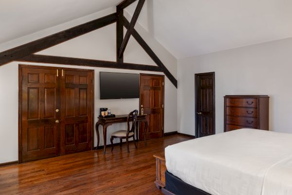 A bedroom with wooden floors, a bed, a dresser, a TV on the wall, a small desk with a chair, and exposed wooden beams.