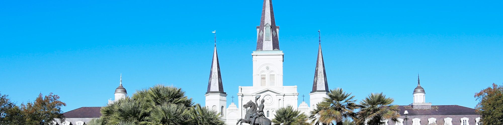 The image shows a park with an equestrian statue, surrounded by trees, in front of a historic building with three spires and a cross.