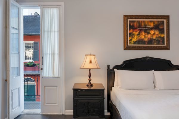 A cozy bedroom with a bed, nightstand, and lamp. There's a painting on the wall and double doors open to a balcony view.