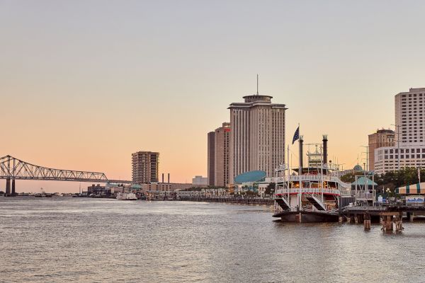 A riverside cityscape with a large bridge, boats on the water, and tall buildings under a clear sky at sunset.