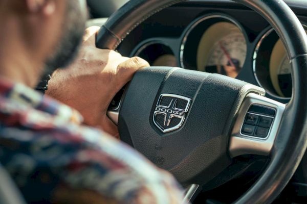 A person is driving a car, holding the steering wheel with a logo visible, focused on the road ahead.