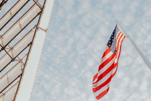 An American flag is waving on a pole against a cloudy sky, with part of a building structure visible on the left side.