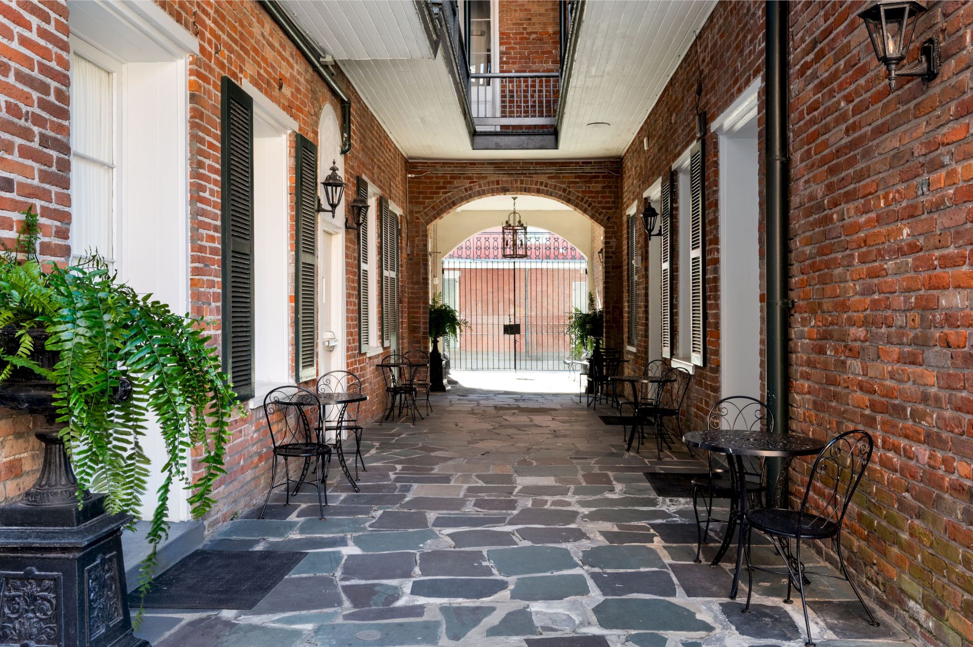 A charming brick-lined passageway with black metal chairs, tables, decorative plants, and lanterns, leading to an ornate gate under an archway.