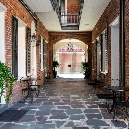 A narrow brick passageway with tables, chairs, ferns, and lanterns leads to a gated courtyard, creating an inviting atmosphere.