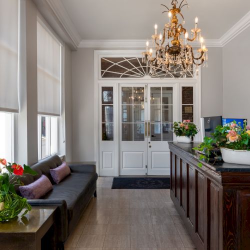 This image shows a bright, elegant lobby with a chandelier, sofa, plants, artwork, and a wooden reception desk by large windows with blinds.