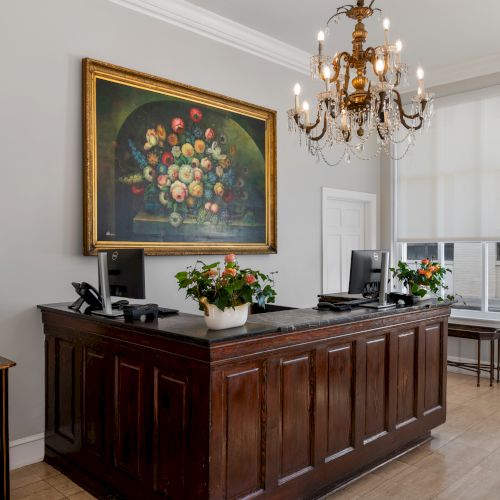 A reception desk with computers, a floral painting, chandelier, and potted plants in an elegantly decorated room.