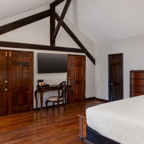 The image shows a bedroom with wooden floors, a bed, dresser, desk, and a mounted TV. Exposed wooden beams are visible on the ceiling.