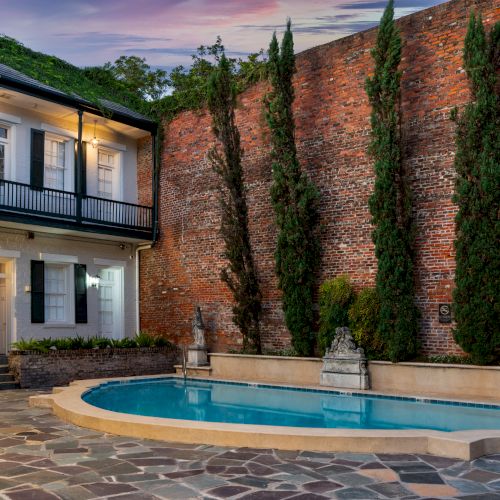 A courtyard with a pool, brick walls, tall trees, and a two-story building with balconies and lights at dusk.