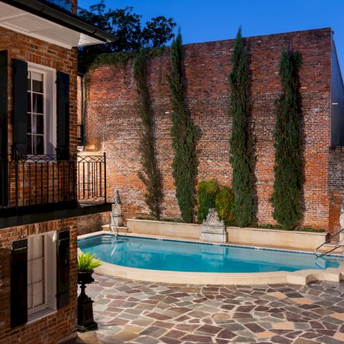 A small, charming courtyard with a swimming pool, brick walls, and tall trees, surrounded by a rustic building.