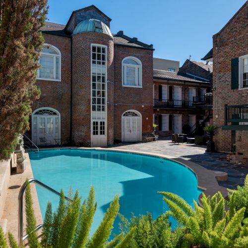 A serene courtyard features a pool surrounded by lush greenery, brick buildings, and a statue, under a clear blue sky.