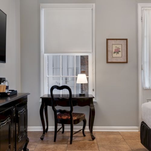 A room with a TV, desk with chair and lamp, a cabinet with a coffee maker, a bed, and a window with blinds.