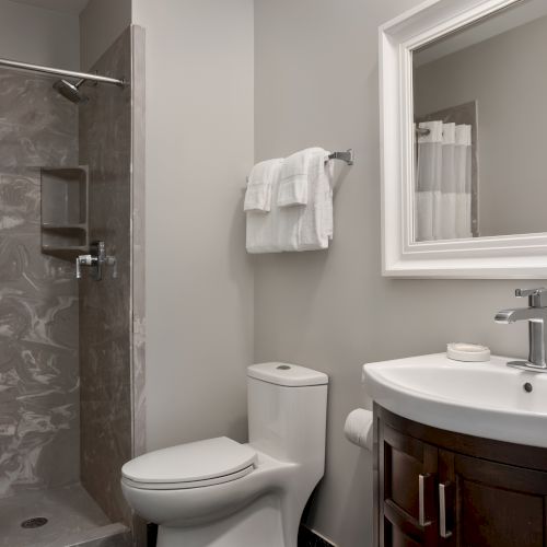 Image of a bathroom with a shower, toilet, and sink with a mirror. Towels are neatly folded on a rack above the toilet.