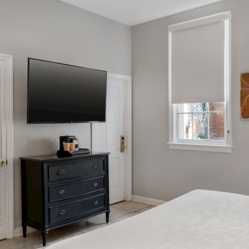 A bedroom with a mounted TV, dresser with coffee maker, window, wall art, bed, and closet door.