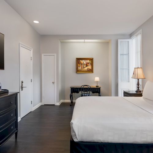 A modern bedroom with a large bed, wall-mounted TV, desk with lamp, dresser, and artwork, featuring dark wood flooring and neutral walls.