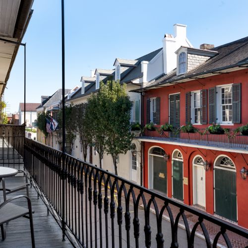 The image shows a balcony view overlooking a street with colorful, historic buildings featuring arched windows and greenery.