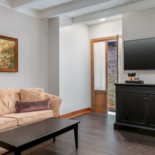 A cozy living room with a beige sofa, painting, black coffee table, TV on a cabinet, and an open door to the outside.