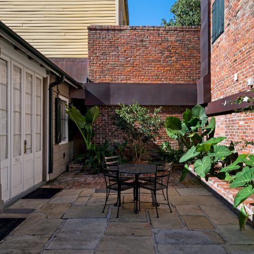 A cozy courtyard with brick walls, leafy plants, and a metal table with chairs on a stone patio.