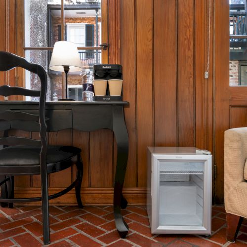 A cozy room with a wooden desk, black chair, lamp, two coffee cups, a white mini-fridge, and a beige armchair on a brick floor.