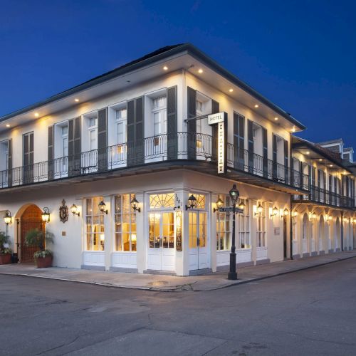 A two-story building with numerous windows and decorative ironwork, lit warmly against an evening sky, positioned on a street corner.