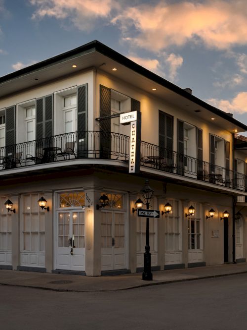 A historic corner building with lit lamps and shuttered windows under a partly cloudy sky, reminiscent of New Orleans architecture.