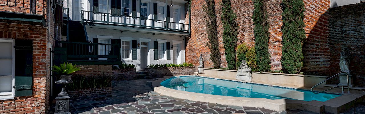 The image shows a courtyard with a small pool and decorative stone flooring, surrounded by a brick wall and trees, adjacent to a building.