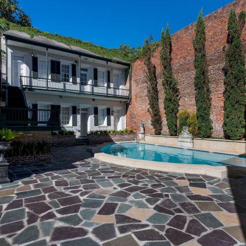 The image shows a courtyard with a small pool and decorative stone flooring, surrounded by a brick wall and trees, adjacent to a building.