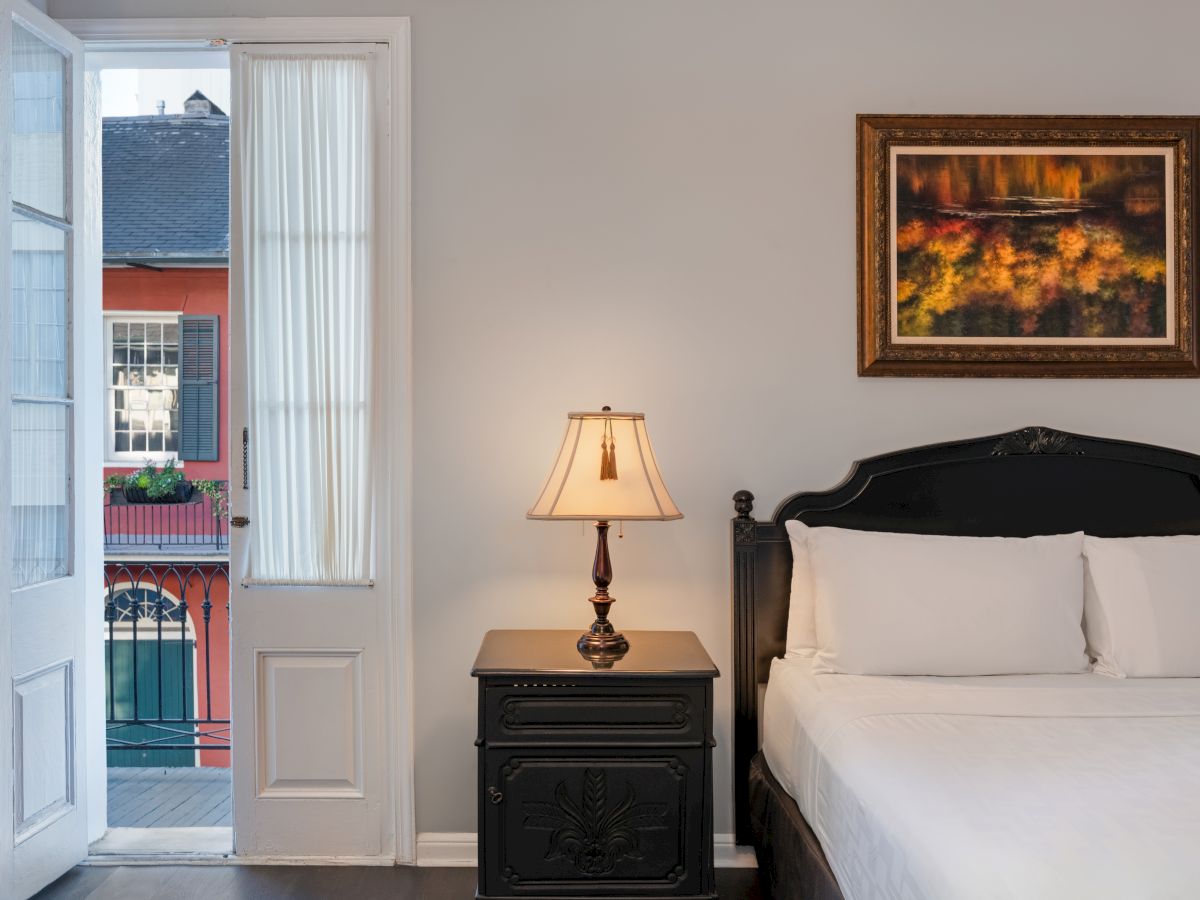 A cozy bedroom with a bed, nightstand, lamp, and painting on the wall, next to a door opening to a view of a colorful building.