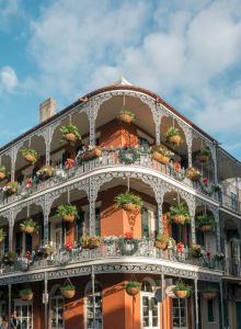 A historic building with ornate ironwork and hanging plants adorns its balconies against a bright blue sky with clouds.