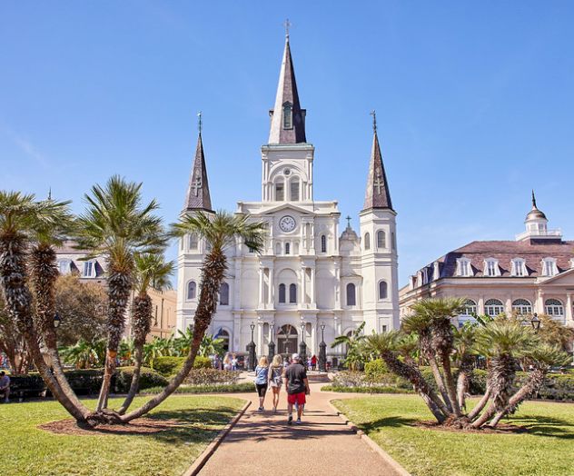 The image shows a historic cathedral with three spires, surrounded by a park with people walking and palm trees in the foreground.