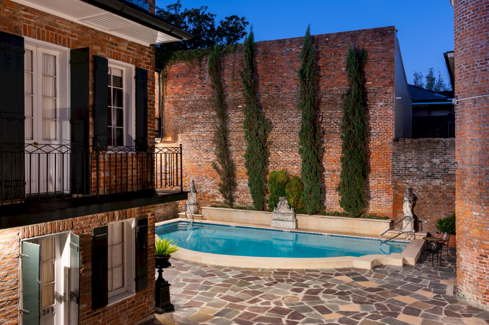 A courtyard with brick walls features a pool, stone statues, and tall cypress trees, surrounded by a stone patio in an evening setting.