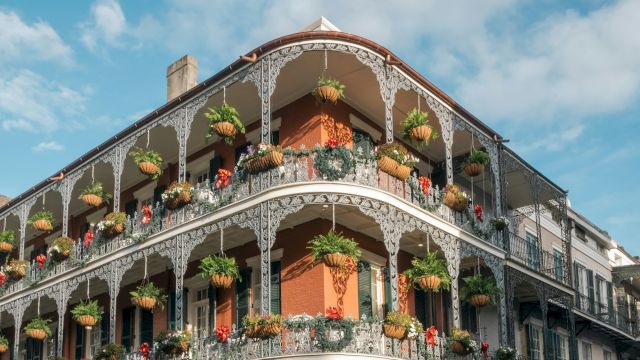 The image features a historic building with ornate ironwork balconies adorned with hanging plants against a partly cloudy sky.