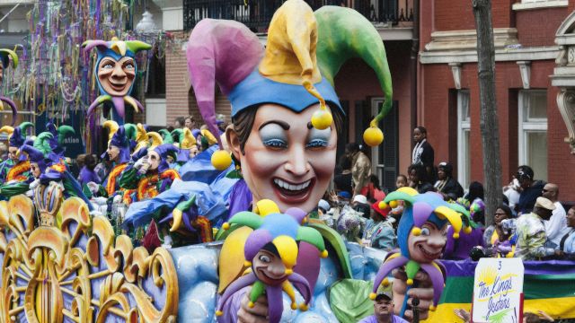 A vibrant Mardi Gras parade float featuring a jester theme, colorful costumes, and a cheerful crowd on a city street.