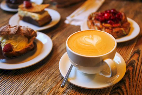 A cappuccino with latte art on a wooden table, surrounded by pastries on plates, including one topped with cherries.