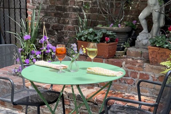 A small garden patio with a green table, two drinks, and a baguette. There's a statue in the background among potted plants.