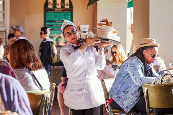 A waiter carries a tray in a busy cafe with seated patrons around, under warm lighting and green awnings.