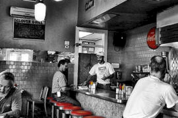 A vintage diner with red stools, black-and-white decor, patrons sitting at the counter, and a server preparing orders in a cozy atmosphere.