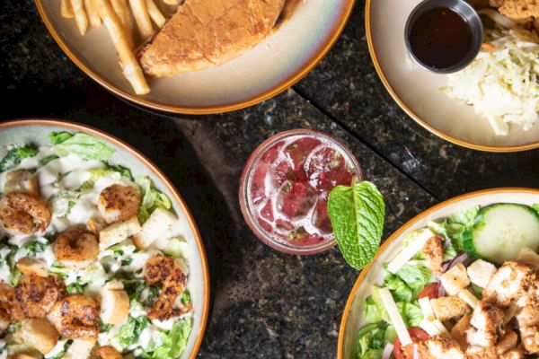 An assortment of dishes including fries with a sandwich, salads with chicken and scallops, and a drink garnished with mint leaves.